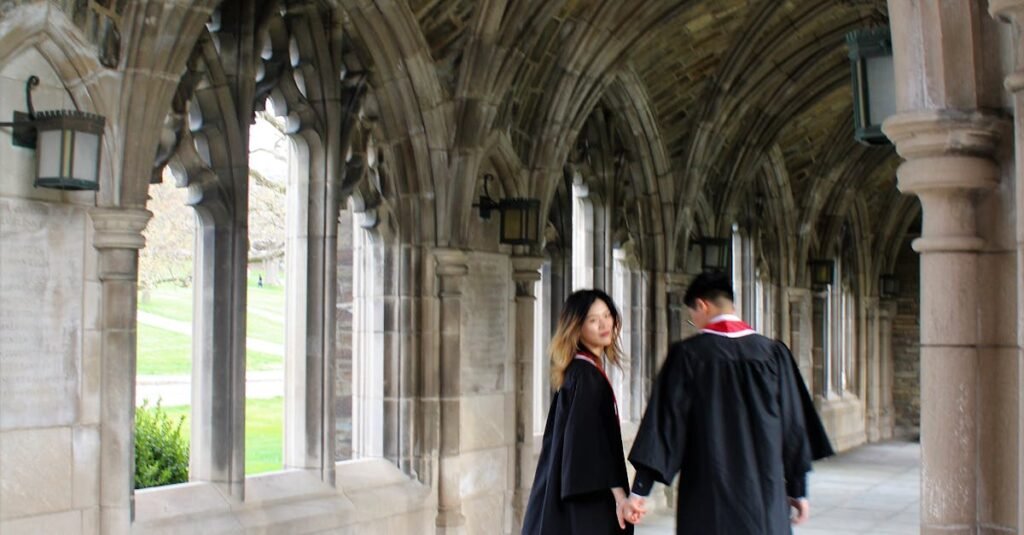 A man and woman in graduation gowns holding hands in a Gothic-style hallway.